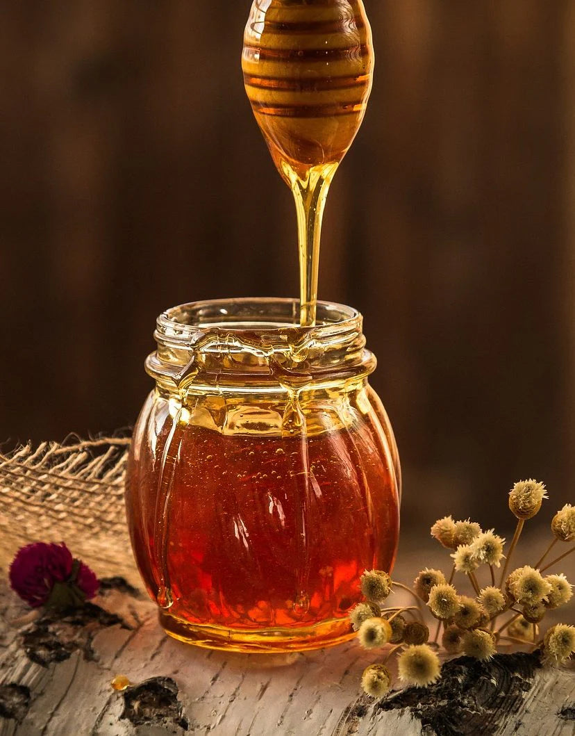 Golden honey drizzling from a wooden dipper into an open glass jar on a wooden surface