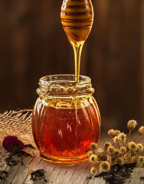 Golden honey drizzling from a wooden dipper into an open glass jar on a wooden surface